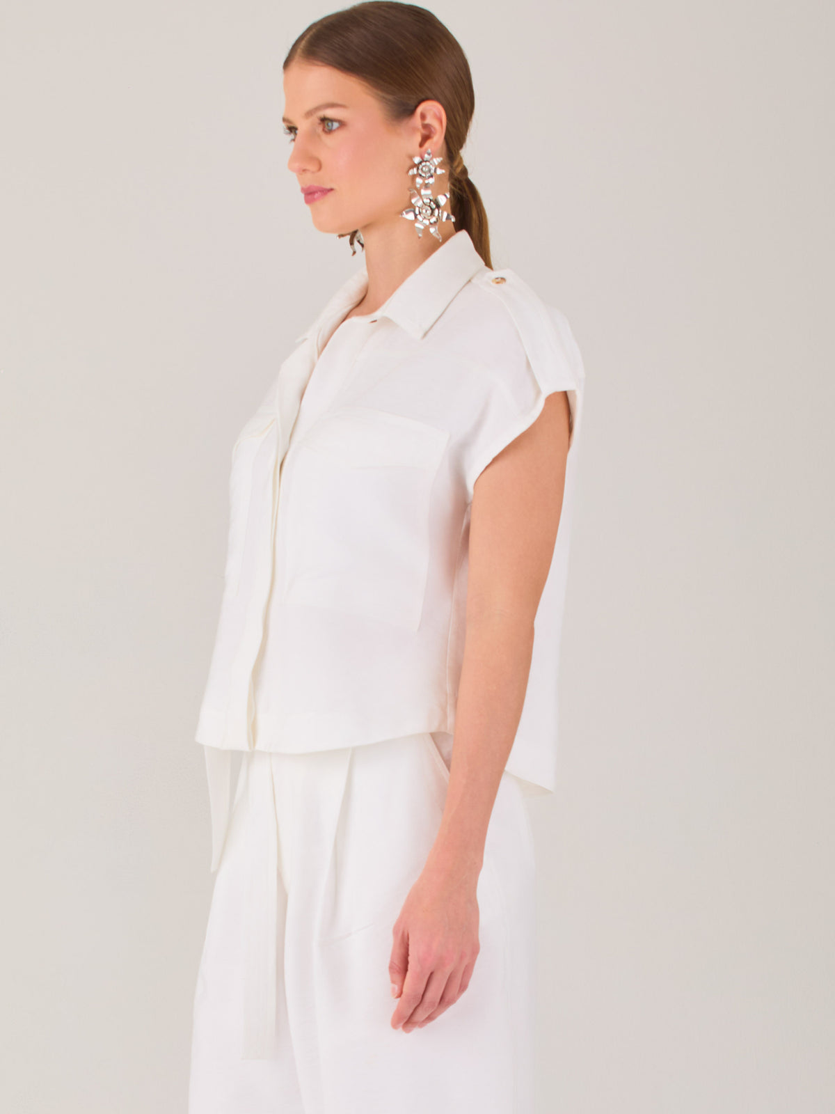 Blusa blanca de estilo camisero con un diseño moderno y minimalista. Cuenta con cuello clásico, tapeta oculta para los botones y mangas cortas tipo caída con detalle de charreteras en los hombros. Posee dos bolsillos de parche en el pecho que le aportan un toque utilitario y estructurado. Su corte recto y fluido la hace ideal tanto para looks casuales como elegantes