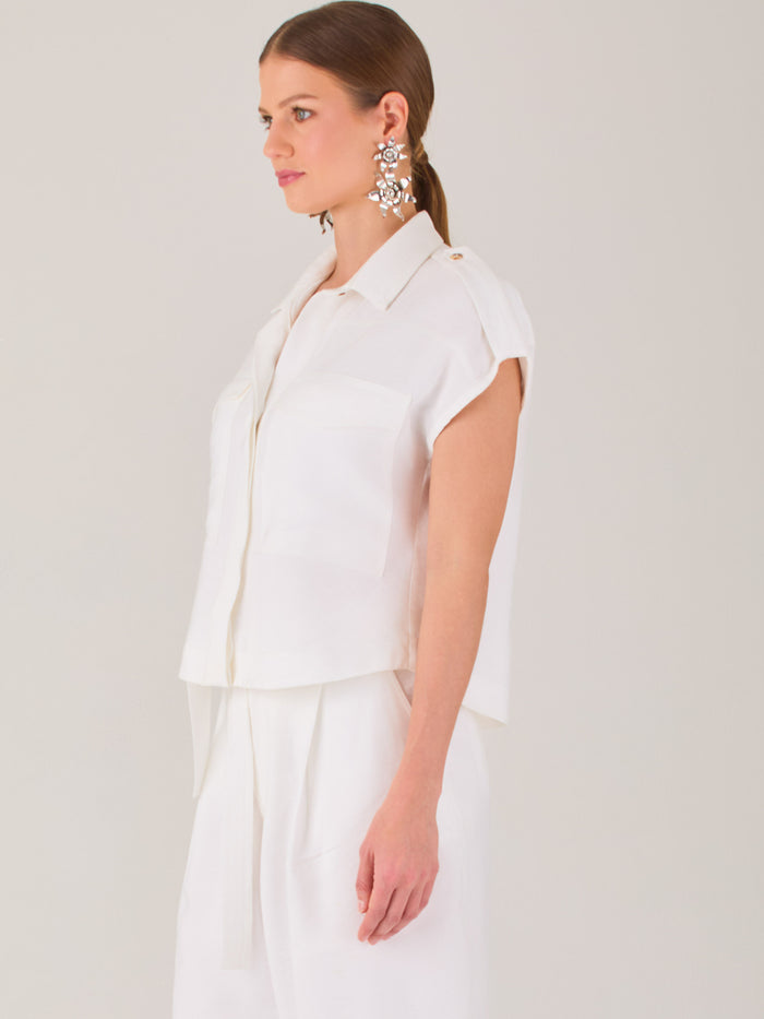 Blusa blanca de estilo camisero con un diseño moderno y minimalista. Cuenta con cuello clásico, tapeta oculta para los botones y mangas cortas tipo caída con detalle de charreteras en los hombros. Posee dos bolsillos de parche en el pecho que le aportan un toque utilitario y estructurado. Su corte recto y fluido la hace ideal tanto para looks casuales como elegantes
