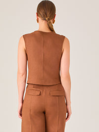 Esta elegante blusa sin mangas presenta un diseño estructurado tipo chaleco, confeccionada en un tejido de textura firme que aporta un aire sofisticado. El tono marrón chocolate refuerza su estilo clásico y versátil. Cuenta con un escote redondo cerrado y una botonadura central con botones metálicos de acabado dorado que destacan sobre el fondo oscuro.