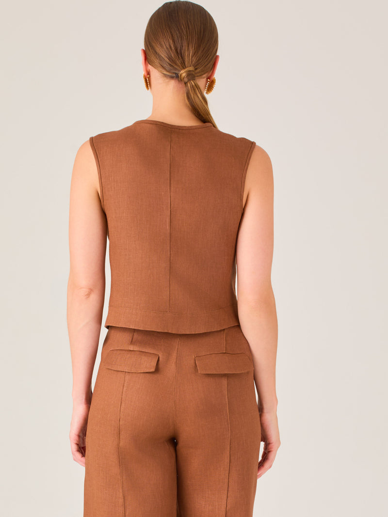Esta elegante blusa sin mangas presenta un diseño estructurado tipo chaleco, confeccionada en un tejido de textura firme que aporta un aire sofisticado. El tono marrón chocolate refuerza su estilo clásico y versátil. Cuenta con un escote redondo cerrado y una botonadura central con botones metálicos de acabado dorado que destacan sobre el fondo oscuro.