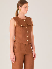 Esta elegante blusa sin mangas presenta un diseño estructurado tipo chaleco, confeccionada en un tejido de textura firme que aporta un aire sofisticado. El tono marrón chocolate refuerza su estilo clásico y versátil. Cuenta con un escote redondo cerrado y una botonadura central con botones metálicos de acabado dorado que destacan sobre el fondo oscuro.