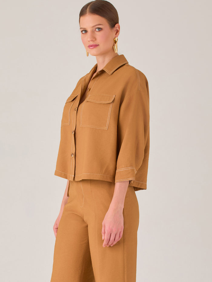 Esta blusa presenta un diseño tipo chaqueta corta (cropped) en un tono beige neutro, ideal para un look sofisticado y contemporáneo. Está confeccionada en una tela estructurada que mantiene su forma, aportando un aire pulido al conjunto. Tiene cuello camisero clásico, cierre frontal con botones metálicos visibles, y dos bolsillos de parche en el pecho con solapa y botón decorativo, que refuerzan el estilo utilitario.

Las mangas son tres cuartos, con un sutil dobladillo al final, lo que le da un toque casua