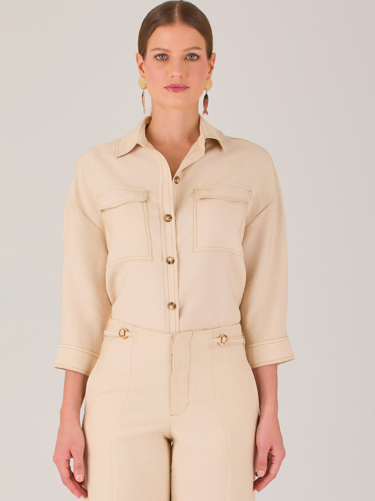 Esta blusa presenta un diseño tipo chaqueta corta (cropped) en un tono beige neutro, ideal para un look sofisticado y contemporáneo. Está confeccionada en una tela estructurada que mantiene su forma, aportando un aire pulido al conjunto. Tiene cuello camisero clásico, cierre frontal con botones metálicos visibles, y dos bolsillos de parche en el pecho con solapa y botón decorativo, que refuerzan el estilo utilitario.
