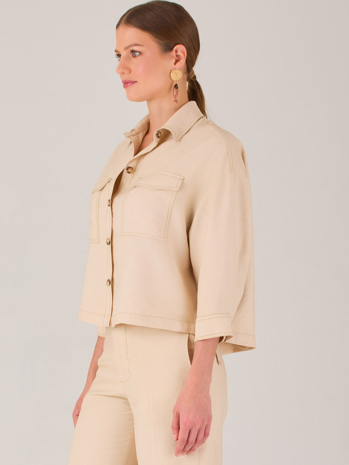 Esta blusa presenta un diseño tipo chaqueta corta (cropped) en un tono beige neutro, ideal para un look sofisticado y contemporáneo. Está confeccionada en una tela estructurada que mantiene su forma, aportando un aire pulido al conjunto. Tiene cuello camisero clásico, cierre frontal con botones metálicos visibles, y dos bolsillos de parche en el pecho con solapa y botón decorativo, que refuerzan el estilo utilitario.
