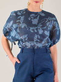 Blusa de manga corta con ajuste elástico en las mangas, corte recto y tejido semitransparente con diseño floral en relieve. Estilo elegante y ligero.