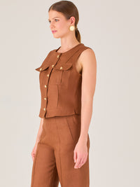 Esta elegante blusa sin mangas presenta un diseño estructurado tipo chaleco, confeccionada en un tejido de textura firme que aporta un aire sofisticado. El tono marrón chocolate refuerza su estilo clásico y versátil. Cuenta con un escote redondo cerrado y una botonadura central con botones metálicos de acabado dorado que destacan sobre el fondo oscuro.