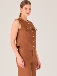 Esta elegante blusa sin mangas presenta un diseño estructurado tipo chaleco, confeccionada en un tejido de textura firme que aporta un aire sofisticado. El tono marrón chocolate refuerza su estilo clásico y versátil. Cuenta con un escote redondo cerrado y una botonadura central con botones metálicos de acabado dorado que destacan sobre el fondo oscuro.