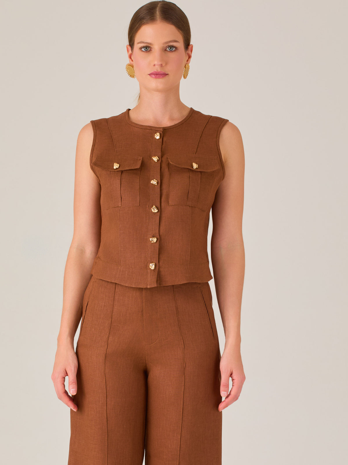 Esta elegante blusa sin mangas presenta un diseño estructurado tipo chaleco, confeccionada en un tejido de textura firme que aporta un aire sofisticado. El tono marrón chocolate refuerza su estilo clásico y versátil. Cuenta con un escote redondo cerrado y una botonadura central con botones metálicos de acabado dorado que destacan sobre el fondo oscuro.