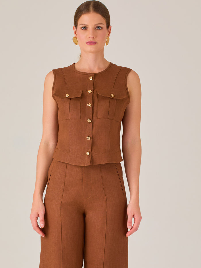 Esta elegante blusa sin mangas presenta un diseño estructurado tipo chaleco, confeccionada en un tejido de textura firme que aporta un aire sofisticado. El tono marrón chocolate refuerza su estilo clásico y versátil. Cuenta con un escote redondo cerrado y una botonadura central con botones metálicos de acabado dorado que destacan sobre el fondo oscuro.