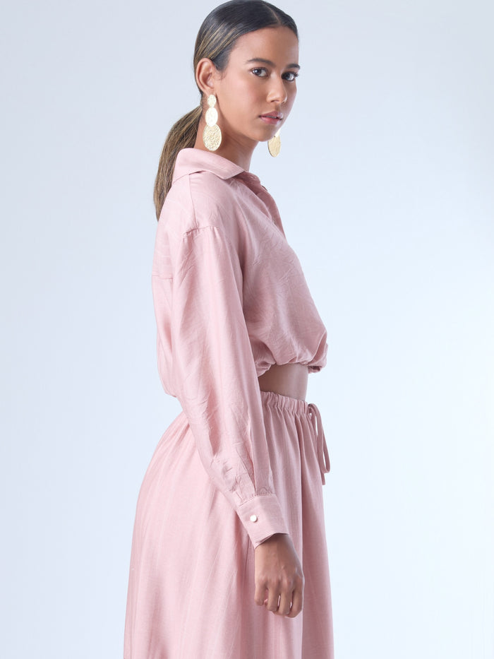 Blusa de corte relajado y largo cropped, con manga larga y puños con botones. Tiene cuello clásico camisero y una botonadura frontal. La prenda se ajusta a la cintura mediante un ligero fruncido con cordón interno, creando un efecto ablusado. Su estilo es casual con un toque sofisticado y femenino.

Detalles:

Color: Rosa