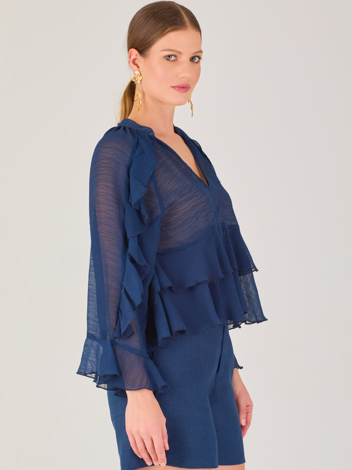 Ligera y romántica, esta blusa de textura vaporosa destaca por sus volantes en capas que aportan movimiento y fluidez. El escote en V y las mangas amplias con acabado ondulado añaden un toque delicado y sofisticado. Ideal para ocasiones especiales, cenas o estilismos casuales con un aire femenino y refinado.