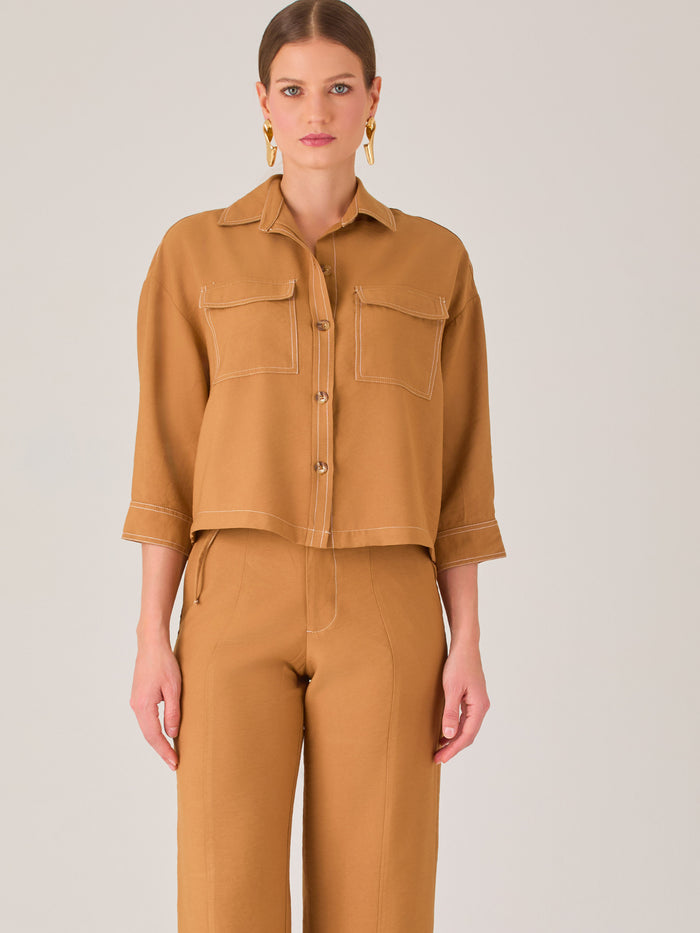 Esta blusa presenta un diseño tipo chaqueta corta (cropped) en un tono beige neutro, ideal para un look sofisticado y contemporáneo. Está confeccionada en una tela estructurada que mantiene su forma, aportando un aire pulido al conjunto. Tiene cuello camisero clásico, cierre frontal con botones metálicos visibles, y dos bolsillos de parche en el pecho con solapa y botón decorativo, que refuerzan el estilo utilitario.

Las mangas son tres cuartos, con un sutil dobladillo al final, lo que le da un toque casua