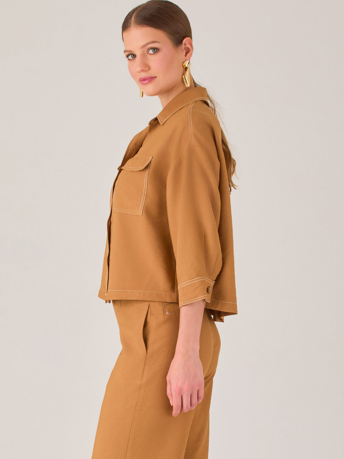 Esta blusa presenta un diseño tipo chaqueta corta (cropped) en un tono beige neutro, ideal para un look sofisticado y contemporáneo. Está confeccionada en una tela estructurada que mantiene su forma, aportando un aire pulido al conjunto. Tiene cuello camisero clásico, cierre frontal con botones metálicos visibles, y dos bolsillos de parche en el pecho con solapa y botón decorativo, que refuerzan el estilo utilitario.

Las mangas son tres cuartos, con un sutil dobladillo al final, lo que le da un toque casua