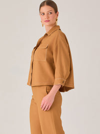 Esta blusa presenta un diseño tipo chaqueta corta (cropped) en un tono beige neutro, ideal para un look sofisticado y contemporáneo. Está confeccionada en una tela estructurada que mantiene su forma, aportando un aire pulido al conjunto. Tiene cuello camisero clásico, cierre frontal con botones metálicos visibles, y dos bolsillos de parche en el pecho con solapa y botón decorativo, que refuerzan el estilo utilitario.

Las mangas son tres cuartos, con un sutil dobladillo al final, lo que le da un toque casua