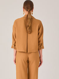 Esta blusa presenta un diseño tipo chaqueta corta (cropped) en un tono beige neutro, ideal para un look sofisticado y contemporáneo. Está confeccionada en una tela estructurada que mantiene su forma, aportando un aire pulido al conjunto. Tiene cuello camisero clásico, cierre frontal con botones metálicos visibles, y dos bolsillos de parche en el pecho con solapa y botón decorativo, que refuerzan el estilo utilitario.

Las mangas son tres cuartos, con un sutil dobladillo al final, lo que le da un toque casua