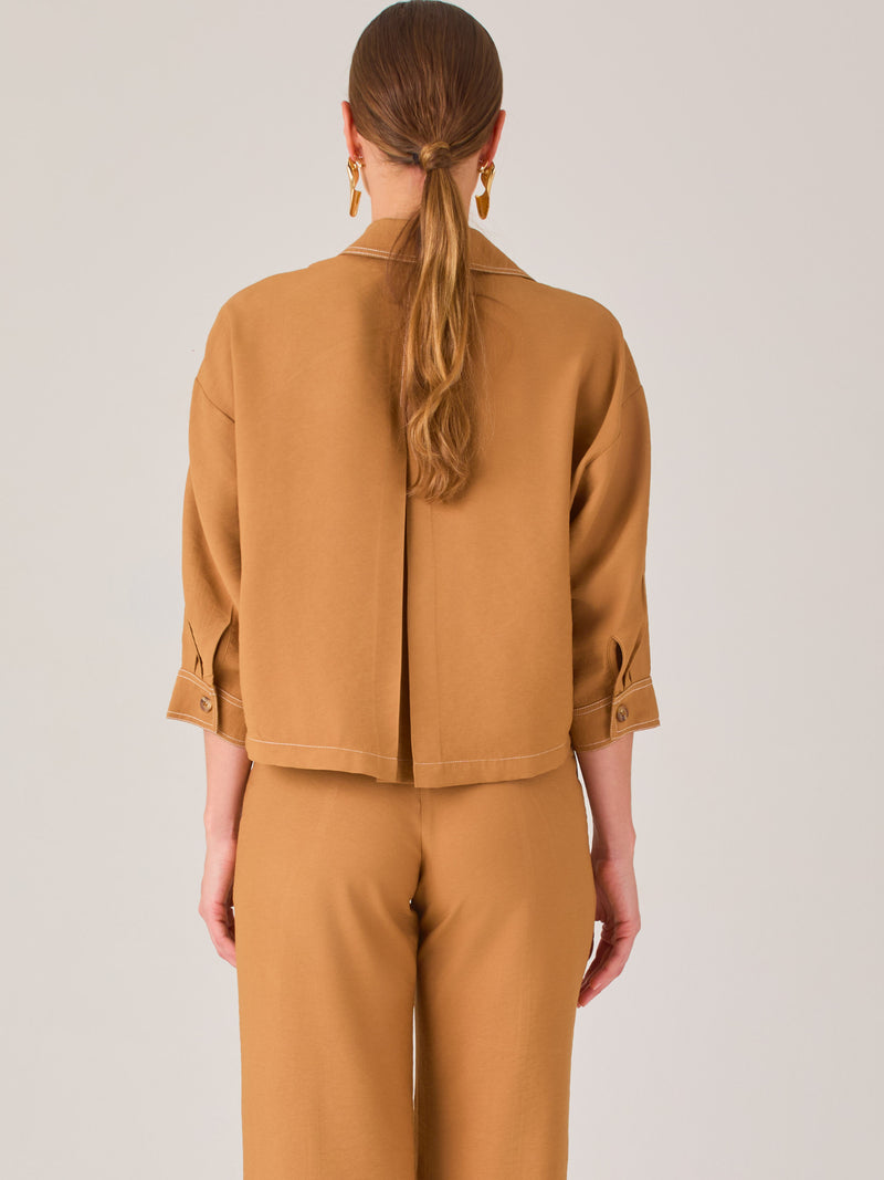 Esta blusa presenta un diseño tipo chaqueta corta (cropped) en un tono beige neutro, ideal para un look sofisticado y contemporáneo. Está confeccionada en una tela estructurada que mantiene su forma, aportando un aire pulido al conjunto. Tiene cuello camisero clásico, cierre frontal con botones metálicos visibles, y dos bolsillos de parche en el pecho con solapa y botón decorativo, que refuerzan el estilo utilitario.

Las mangas son tres cuartos, con un sutil dobladillo al final, lo que le da un toque casua