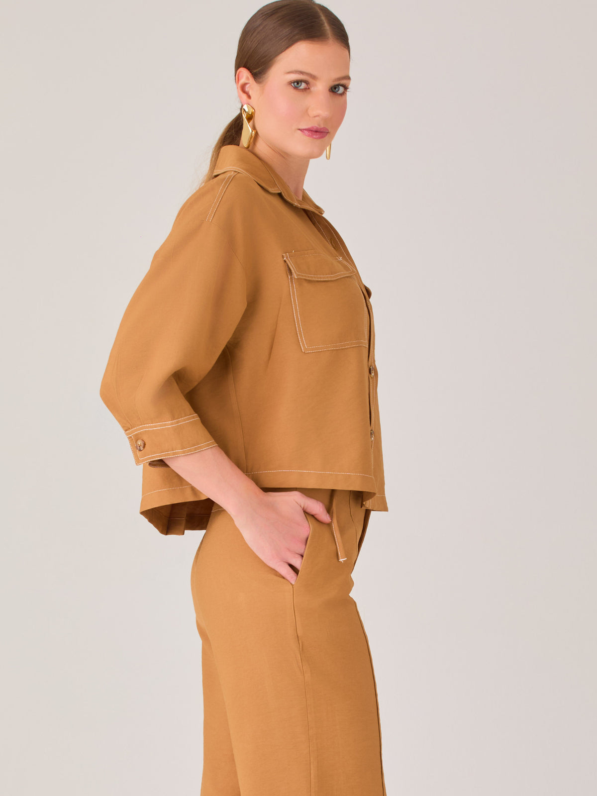 Esta blusa presenta un diseño tipo chaqueta corta (cropped) en un tono beige neutro, ideal para un look sofisticado y contemporáneo. Está confeccionada en una tela estructurada que mantiene su forma, aportando un aire pulido al conjunto. Tiene cuello camisero clásico, cierre frontal con botones metálicos visibles, y dos bolsillos de parche en el pecho con solapa y botón decorativo, que refuerzan el estilo utilitario.

Las mangas son tres cuartos, con un sutil dobladillo al final, lo que le da un toque casua
