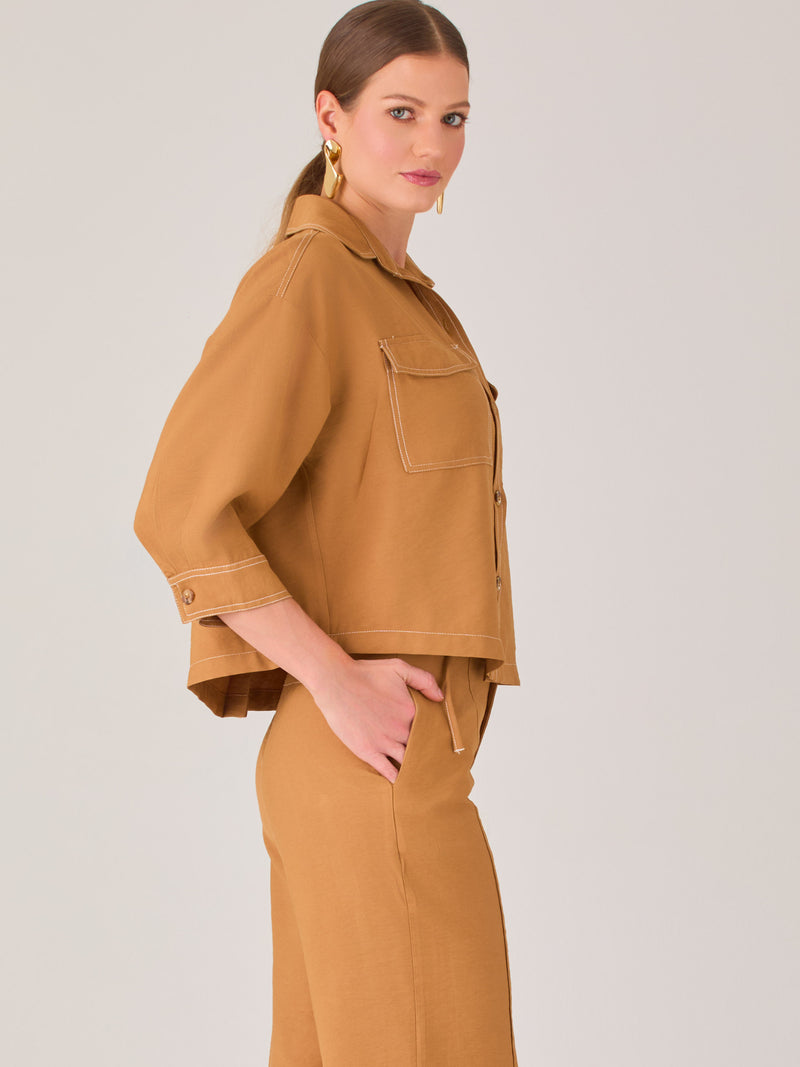 Esta blusa presenta un diseño tipo chaqueta corta (cropped) en un tono beige neutro, ideal para un look sofisticado y contemporáneo. Está confeccionada en una tela estructurada que mantiene su forma, aportando un aire pulido al conjunto. Tiene cuello camisero clásico, cierre frontal con botones metálicos visibles, y dos bolsillos de parche en el pecho con solapa y botón decorativo, que refuerzan el estilo utilitario.

Las mangas son tres cuartos, con un sutil dobladillo al final, lo que le da un toque casua