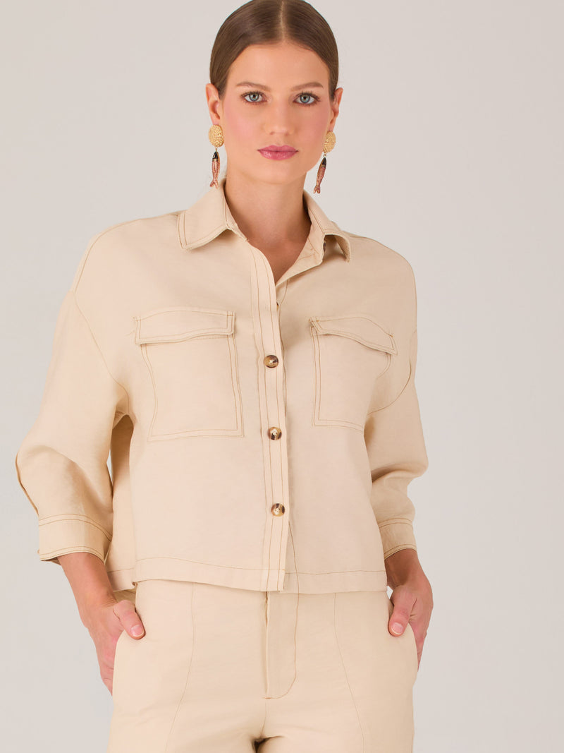 Esta blusa presenta un diseño tipo chaqueta corta (cropped) en un tono beige neutro, ideal para un look sofisticado y contemporáneo. Está confeccionada en una tela estructurada que mantiene su forma, aportando un aire pulido al conjunto. Tiene cuello camisero clásico, cierre frontal con botones metálicos visibles, y dos bolsillos de parche en el pecho con solapa y botón decorativo, que refuerzan el estilo utilitario.
