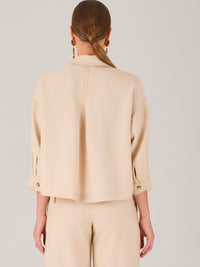 Esta blusa presenta un diseño tipo chaqueta corta (cropped) en un tono beige neutro, ideal para un look sofisticado y contemporáneo. Está confeccionada en una tela estructurada que mantiene su forma, aportando un aire pulido al conjunto. Tiene cuello camisero clásico, cierre frontal con botones metálicos visibles, y dos bolsillos de parche en el pecho con solapa y botón decorativo, que refuerzan el estilo utilitario.