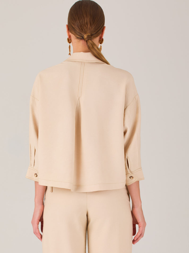 Esta blusa presenta un diseño tipo chaqueta corta (cropped) en un tono beige neutro, ideal para un look sofisticado y contemporáneo. Está confeccionada en una tela estructurada que mantiene su forma, aportando un aire pulido al conjunto. Tiene cuello camisero clásico, cierre frontal con botones metálicos visibles, y dos bolsillos de parche en el pecho con solapa y botón decorativo, que refuerzan el estilo utilitario.