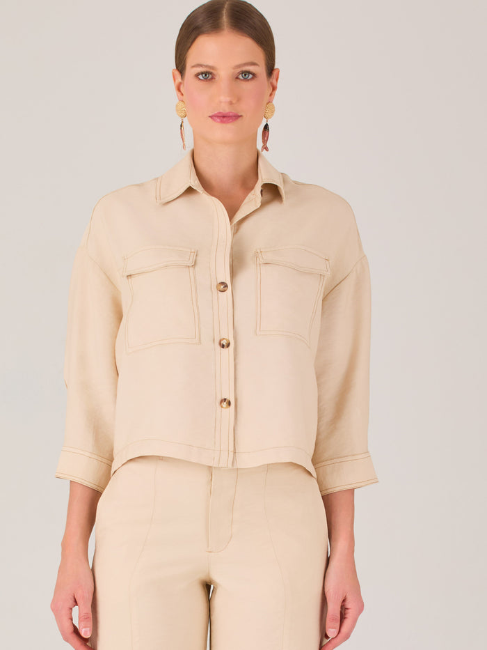 Esta blusa presenta un diseño tipo chaqueta corta (cropped) en un tono beige neutro, ideal para un look sofisticado y contemporáneo. Está confeccionada en una tela estructurada que mantiene su forma, aportando un aire pulido al conjunto. Tiene cuello camisero clásico, cierre frontal con botones metálicos visibles, y dos bolsillos de parche en el pecho con solapa y botón decorativo, que refuerzan el estilo utilitario.