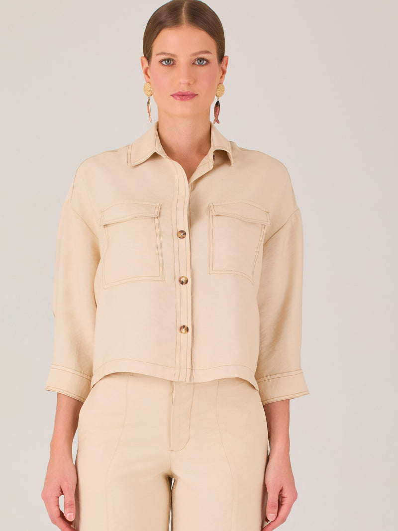 Esta blusa presenta un diseño tipo chaqueta corta (cropped) en un tono beige neutro, ideal para un look sofisticado y contemporáneo. Está confeccionada en una tela estructurada que mantiene su forma, aportando un aire pulido al conjunto. Tiene cuello camisero clásico, cierre frontal con botones metálicos visibles, y dos bolsillos de parche en el pecho con solapa y botón decorativo, que refuerzan el estilo utilitario.