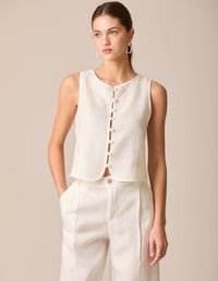 Top tipo chaleco en lino blanco perla, sin mangas y con escote redondo. Cierre frontal con botones forrados y diseño entallado en la parte superior. Su dobladillo curvado aporta un toque estructurado y sofisticado, ideal para outfits frescos y elegantes.