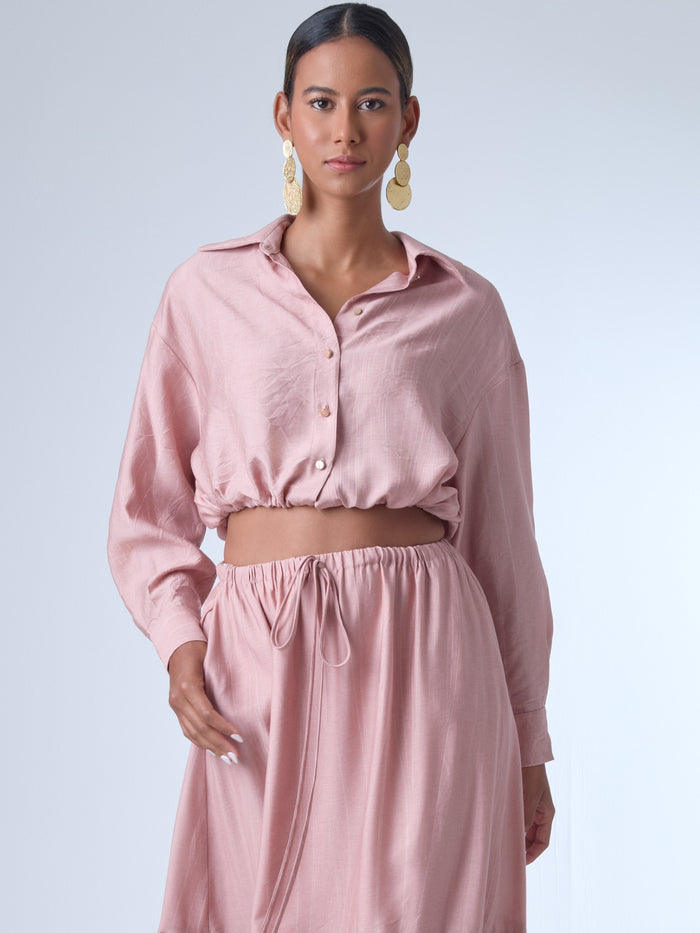 Blusa de corte relajado y largo cropped, con manga larga y puños con botones. Tiene cuello clásico camisero y una botonadura frontal. La prenda se ajusta a la cintura mediante un ligero fruncido con cordón interno, creando un efecto ablusado. Su estilo es casual con un toque sofisticado y femenino.

Detalles:

Color: Rosa