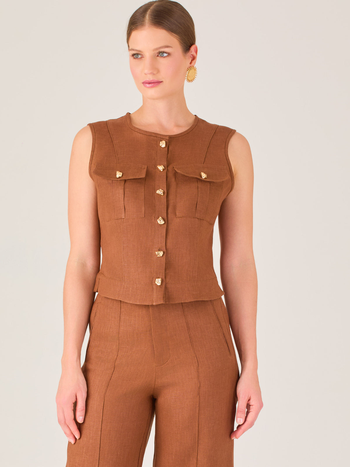 Esta elegante blusa sin mangas presenta un diseño estructurado tipo chaleco, confeccionada en un tejido de textura firme que aporta un aire sofisticado. El tono marrón chocolate refuerza su estilo clásico y versátil. Cuenta con un escote redondo cerrado y una botonadura central con botones metálicos de acabado dorado que destacan sobre el fondo oscuro.