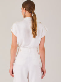 Blusa blanca de estilo camisero con un diseño moderno y minimalista. Cuenta con cuello clásico, tapeta oculta para los botones y mangas cortas tipo caída con detalle de charreteras en los hombros. Posee dos bolsillos de parche en el pecho que le aportan un toque utilitario y estructurado. Su corte recto y fluido la hace ideal tanto para looks casuales como elegantes