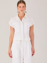 Blusa blanca de estilo camisero con un diseño moderno y minimalista. Cuenta con cuello clásico, tapeta oculta para los botones y mangas cortas tipo caída con detalle de charreteras en los hombros. Posee dos bolsillos de parche en el pecho que le aportan un toque utilitario y estructurado. Su corte recto y fluido la hace ideal tanto para looks casuales como elegantes