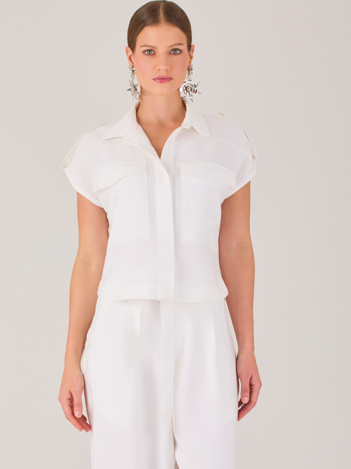 Blusa blanca de estilo camisero con un diseño moderno y minimalista. Cuenta con cuello clásico, tapeta oculta para los botones y mangas cortas tipo caída con detalle de charreteras en los hombros. Posee dos bolsillos de parche en el pecho que le aportan un toque utilitario y estructurado. Su corte recto y fluido la hace ideal tanto para looks casuales como elegantes