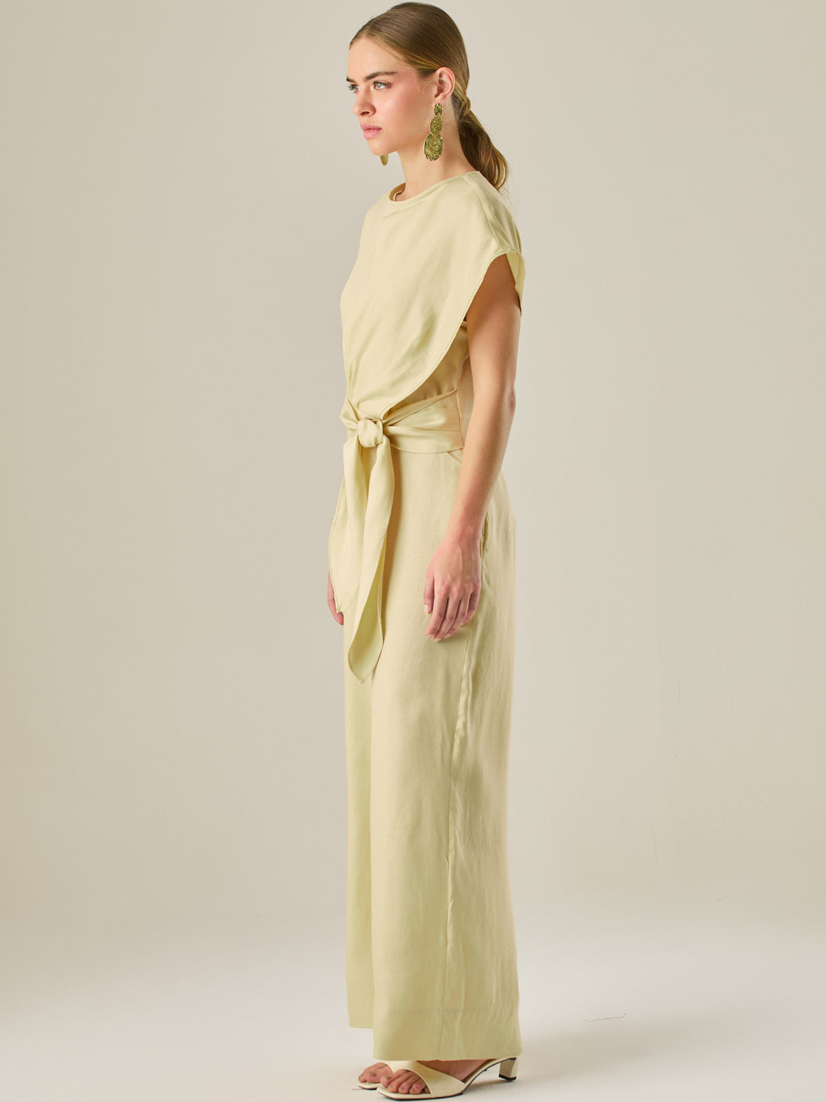 Este enterizo presenta un diseño elegante y minimalista, ideal para ocasiones casuales con estilo o eventos diurnos sofisticados. Confeccionado en un tono beige claro, tiene un corte relajado con mangas tipo kimono, cuello redondo y un diseño limpio sin botones ni cierres visibles en el frente. La prenda se ajusta en la cintura con un cinturón del mismo tejido, que se anuda al frente, creando una silueta favorecedora. El pantalón es de pierna ancha y caída fluida, lo que aporta comodidad y movimiento. Compl