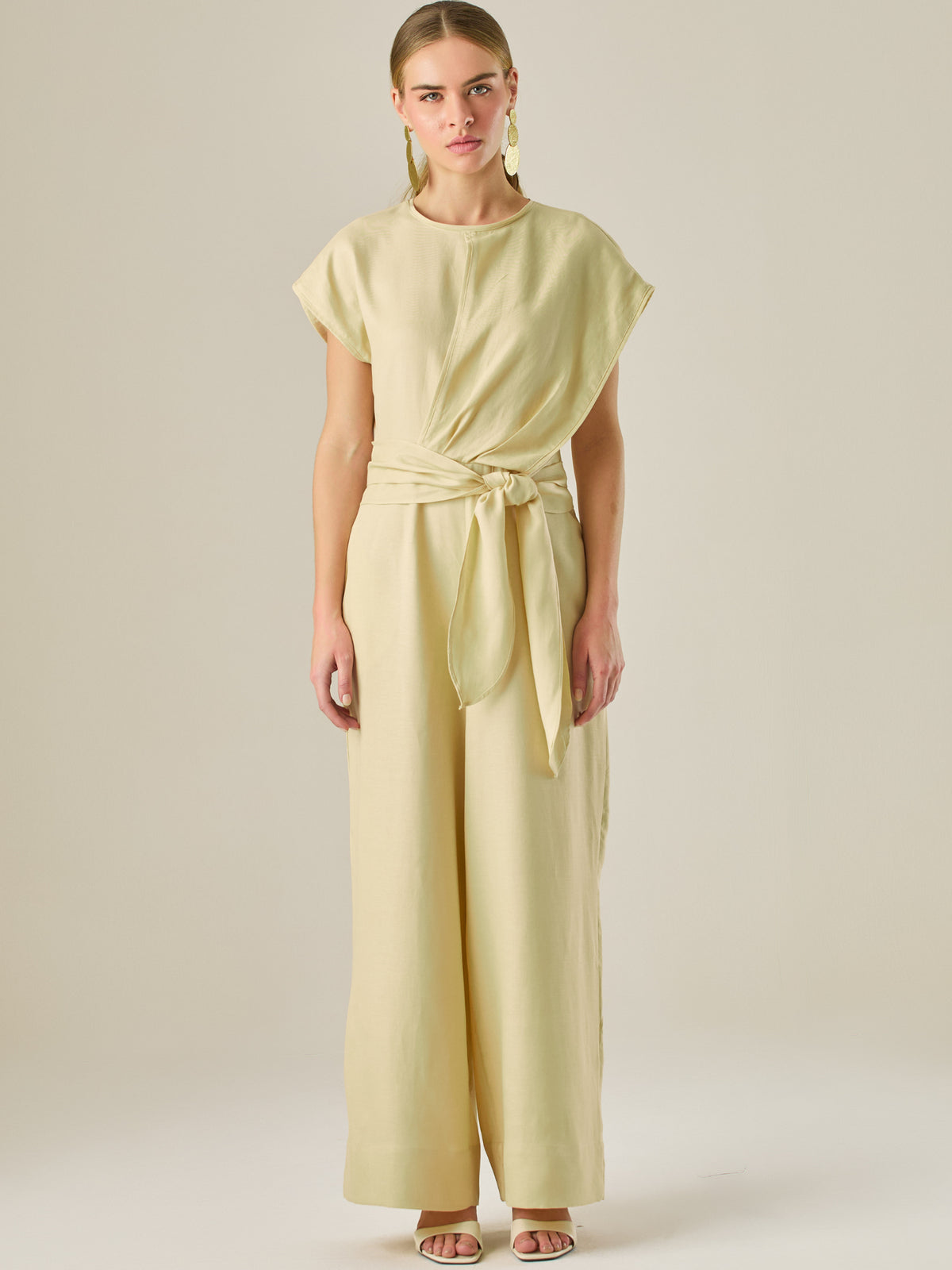 Este enterizo presenta un diseño elegante y minimalista, ideal para ocasiones casuales con estilo o eventos diurnos sofisticados. Confeccionado en un tono beige claro, tiene un corte relajado con mangas tipo kimono, cuello redondo y un diseño limpio sin botones ni cierres visibles en el frente. La prenda se ajusta en la cintura con un cinturón del mismo tejido, que se anuda al frente, creando una silueta favorecedora. El pantalón es de pierna ancha y caída fluida, lo que aporta comodidad y movimiento. Compl