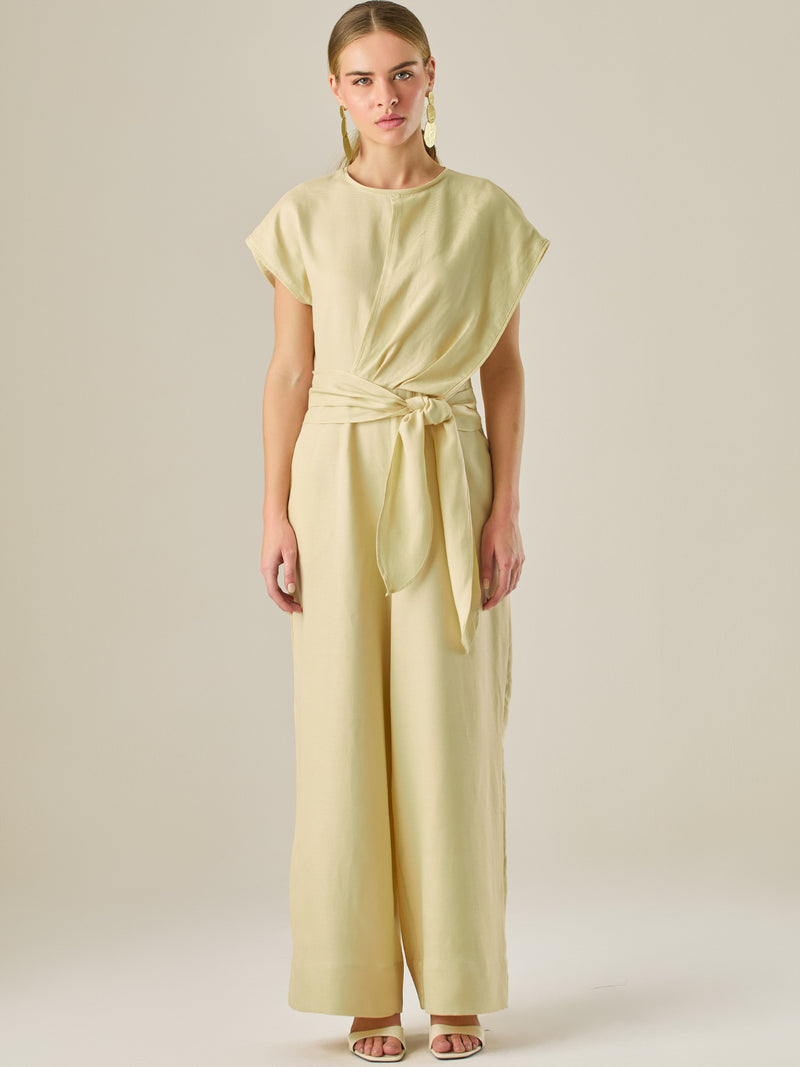 Este enterizo presenta un diseño elegante y minimalista, ideal para ocasiones casuales con estilo o eventos diurnos sofisticados. Confeccionado en un tono beige claro, tiene un corte relajado con mangas tipo kimono, cuello redondo y un diseño limpio sin botones ni cierres visibles en el frente. La prenda se ajusta en la cintura con un cinturón del mismo tejido, que se anuda al frente, creando una silueta favorecedora. El pantalón es de pierna ancha y caída fluida, lo que aporta comodidad y movimiento. Compl