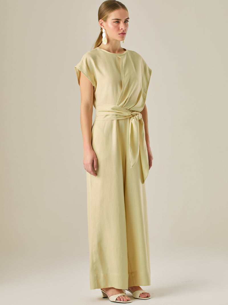 Este enterizo presenta un diseño elegante y minimalista, ideal para ocasiones casuales con estilo o eventos diurnos sofisticados. Confeccionado en un tono beige claro, tiene un corte relajado con mangas tipo kimono, cuello redondo y un diseño limpio sin botones ni cierres visibles en el frente. La prenda se ajusta en la cintura con un cinturón del mismo tejido, que se anuda al frente, creando una silueta favorecedora. El pantalón es de pierna ancha y caída fluida, lo que aporta comodidad y movimiento. Compl