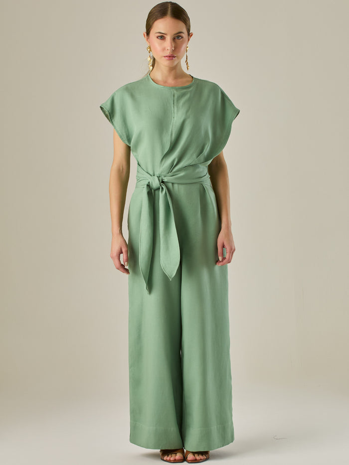 Este enterizo presenta un diseño elegante y minimalista, ideal para ocasiones casuales con estilo o eventos diurnos sofisticados. Confeccionado en un tono beige claro, tiene un corte relajado con mangas tipo kimono, cuello redondo y un diseño limpio sin botones ni cierres visibles en el frente. La prenda se ajusta en la cintura con un cinturón del mismo tejido, que se anuda al frente, creando una silueta favorecedora. El pantalón es de pierna ancha y caída fluida, lo que aporta comodidad y movimiento. Compl