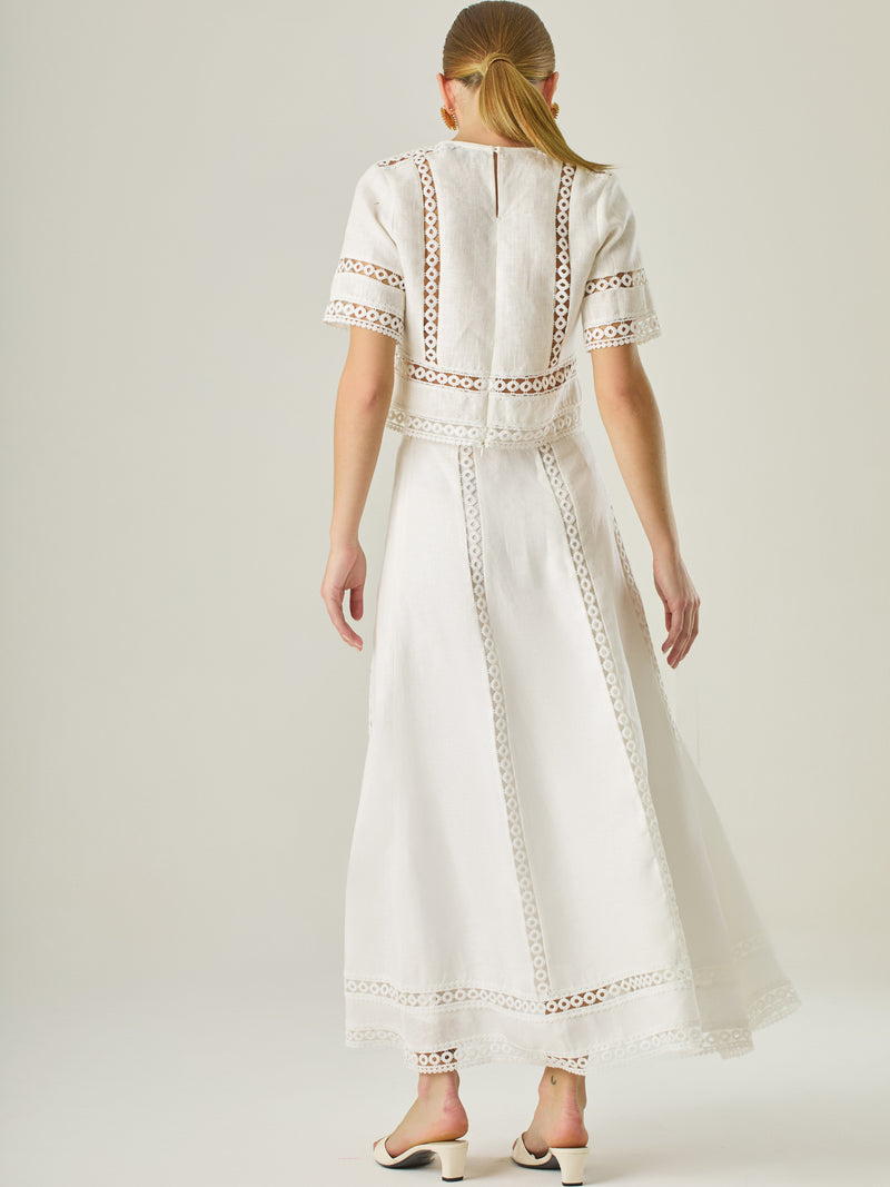 Esta elegante falda larga está confeccionada en un tejido blanco de apariencia ligera y fresca, ideal para climas cálidos o estilos veraniegos. Su diseño de silueta A le da una caída fluida y femenina, que aporta comodidad sin perder sofisticación. La falda destaca por sus aplicaciones de encaje calado en líneas verticales y una franja decorativa en el bajo, que añade un aire romántico y artesanal a la prenda. La cintura alta estiliza la figura y permite combinarla fácilmente con tops por dentro. Es perfect