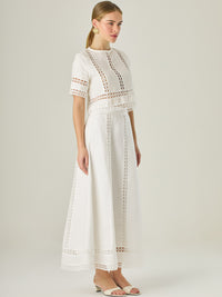 Esta elegante falda larga está confeccionada en un tejido blanco de apariencia ligera y fresca, ideal para climas cálidos o estilos veraniegos. Su diseño de silueta A le da una caída fluida y femenina, que aporta comodidad sin perder sofisticación. La falda destaca por sus aplicaciones de encaje calado en líneas verticales y una franja decorativa en el bajo, que añade un aire romántico y artesanal a la prenda. La cintura alta estiliza la figura y permite combinarla fácilmente con tops por dentro. Es perfect
