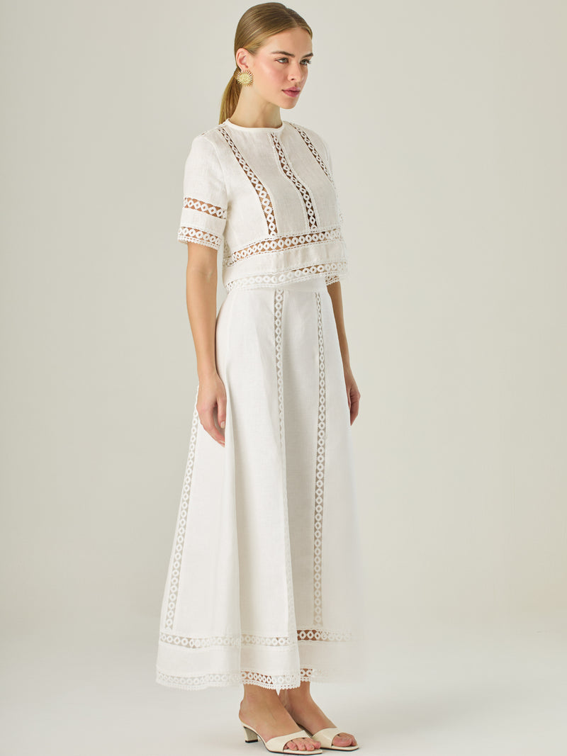 Esta elegante falda larga está confeccionada en un tejido blanco de apariencia ligera y fresca, ideal para climas cálidos o estilos veraniegos. Su diseño de silueta A le da una caída fluida y femenina, que aporta comodidad sin perder sofisticación. La falda destaca por sus aplicaciones de encaje calado en líneas verticales y una franja decorativa en el bajo, que añade un aire romántico y artesanal a la prenda. La cintura alta estiliza la figura y permite combinarla fácilmente con tops por dentro. Es perfect