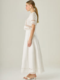 Esta elegante falda larga está confeccionada en un tejido blanco de apariencia ligera y fresca, ideal para climas cálidos o estilos veraniegos. Su diseño de silueta A le da una caída fluida y femenina, que aporta comodidad sin perder sofisticación. La falda destaca por sus aplicaciones de encaje calado en líneas verticales y una franja decorativa en el bajo, que añade un aire romántico y artesanal a la prenda. La cintura alta estiliza la figura y permite combinarla fácilmente con tops por dentro. Es perfect