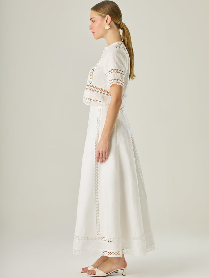 Esta elegante falda larga está confeccionada en un tejido blanco de apariencia ligera y fresca, ideal para climas cálidos o estilos veraniegos. Su diseño de silueta A le da una caída fluida y femenina, que aporta comodidad sin perder sofisticación. La falda destaca por sus aplicaciones de encaje calado en líneas verticales y una franja decorativa en el bajo, que añade un aire romántico y artesanal a la prenda. La cintura alta estiliza la figura y permite combinarla fácilmente con tops por dentro. Es perfect
