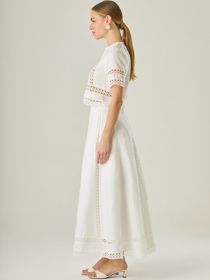 Esta elegante falda larga está confeccionada en un tejido blanco de apariencia ligera y fresca, ideal para climas cálidos o estilos veraniegos. Su diseño de silueta A le da una caída fluida y femenina, que aporta comodidad sin perder sofisticación. La falda destaca por sus aplicaciones de encaje calado en líneas verticales y una franja decorativa en el bajo, que añade un aire romántico y artesanal a la prenda. La cintura alta estiliza la figura y permite combinarla fácilmente con tops por dentro. Es perfect