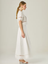 Esta elegante falda larga está confeccionada en un tejido blanco de apariencia ligera y fresca, ideal para climas cálidos o estilos veraniegos. Su diseño de silueta A le da una caída fluida y femenina, que aporta comodidad sin perder sofisticación. La falda destaca por sus aplicaciones de encaje calado en líneas verticales y una franja decorativa en el bajo, que añade un aire romántico y artesanal a la prenda. La cintura alta estiliza la figura y permite combinarla fácilmente con tops por dentro. Es perfect