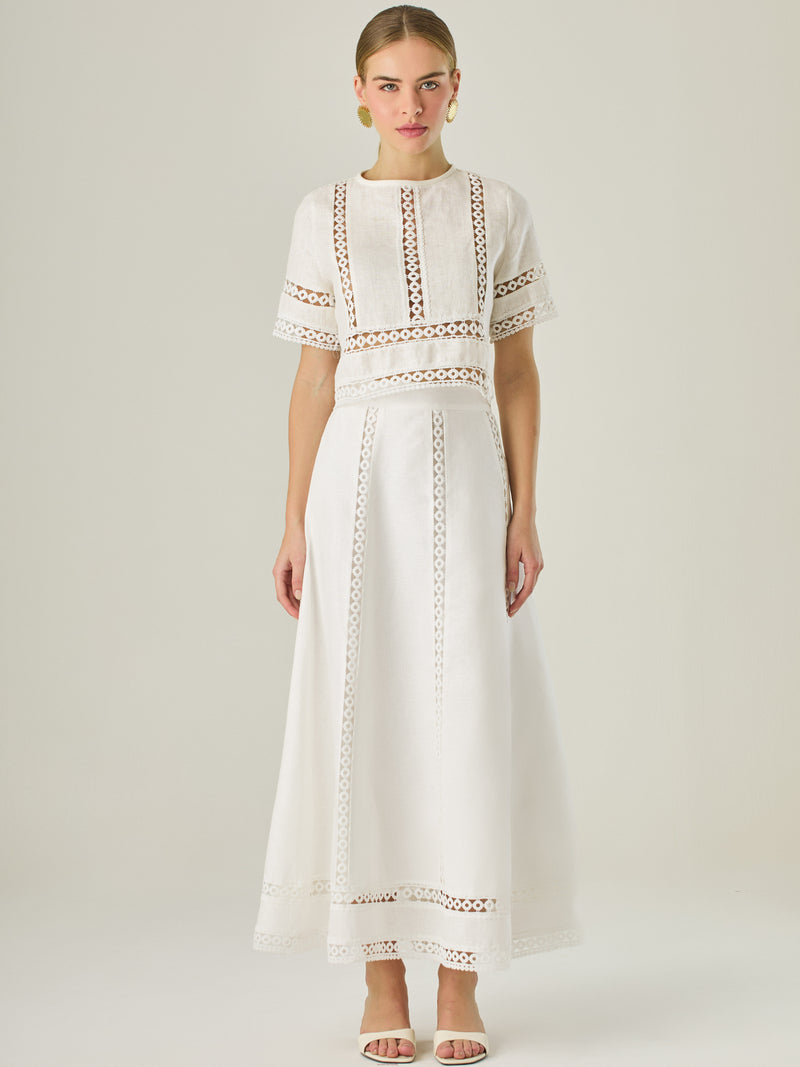 Esta elegante falda larga está confeccionada en un tejido blanco de apariencia ligera y fresca, ideal para climas cálidos o estilos veraniegos. Su diseño de silueta A le da una caída fluida y femenina, que aporta comodidad sin perder sofisticación. La falda destaca por sus aplicaciones de encaje calado en líneas verticales y una franja decorativa en el bajo, que añade un aire romántico y artesanal a la prenda. La cintura alta estiliza la figura y permite combinarla fácilmente con tops por dentro. Es perfect