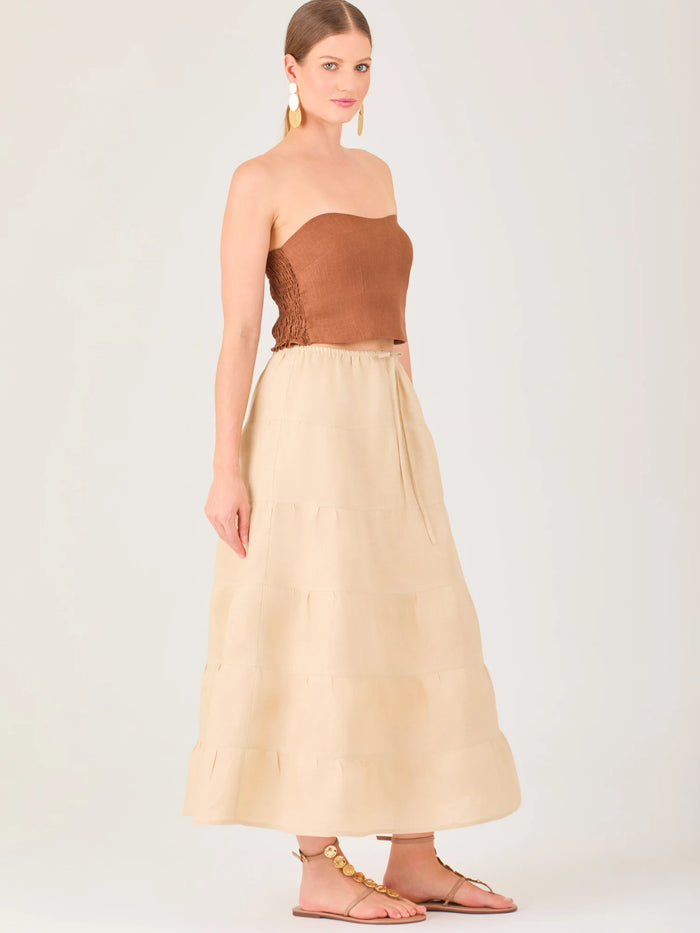 La falda es de largo midi, confeccionada en una tela ligera de tono beige claro. Presenta un diseño de corte amplio con caída fluida y detalles de costuras horizontales que crean un efecto de franjas sutiles a lo largo de la prenda. La pretina es elástica con cordón ajustable al frente, lo que le aporta comodidad y un estilo relajado. Su diseño minimalista y neutral la hace versátil, ideal para combinar con diferentes tops y accesorios, manteniendo un aire fresco y natural.