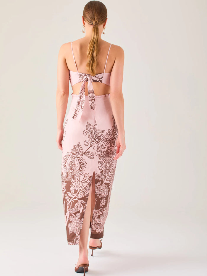 El vestido es largo y de tirantes finos, con escote fruncido y cortes laterales que dejan parte de la cintura descubierta. La falda presenta un estampado tipo floral y paisley en tonos azul profundo sobre fondo blanco, concentrado en la parte baja y difuminándose hacia arriba, creando un contraste elegante y veraniego. El diseño combina frescura y sofisticación, ideal para climas cálidos.