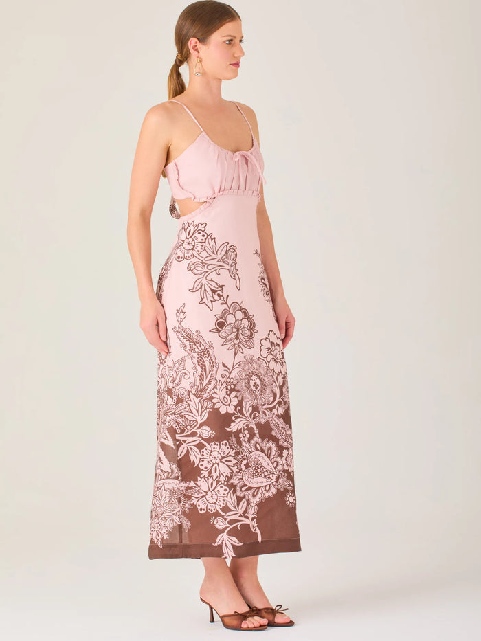 El vestido es largo y de tirantes finos, con escote fruncido y cortes laterales que dejan parte de la cintura descubierta. La falda presenta un estampado tipo floral y paisley en tonos azul profundo sobre fondo blanco, concentrado en la parte baja y difuminándose hacia arriba, creando un contraste elegante y veraniego. El diseño combina frescura y sofisticación, ideal para climas cálidos.