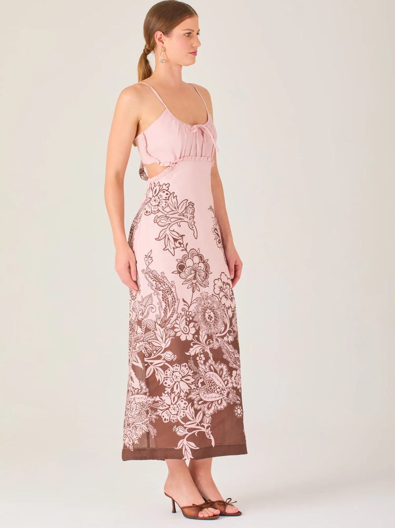 El vestido es largo y de tirantes finos, con escote fruncido y cortes laterales que dejan parte de la cintura descubierta. La falda presenta un estampado tipo floral y paisley en tonos azul profundo sobre fondo blanco, concentrado en la parte baja y difuminándose hacia arriba, creando un contraste elegante y veraniego. El diseño combina frescura y sofisticación, ideal para climas cálidos.